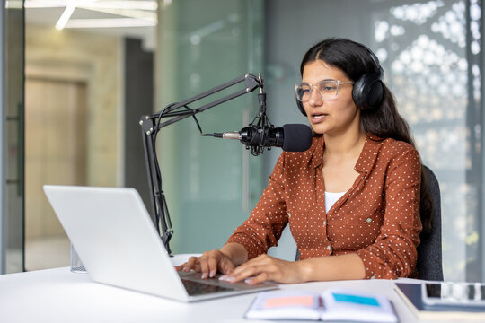 Indian woman wearing headphones and glasses, engaging in a podcast recording session in a modern studio, speaking into a professional microphone while typing on a laptop