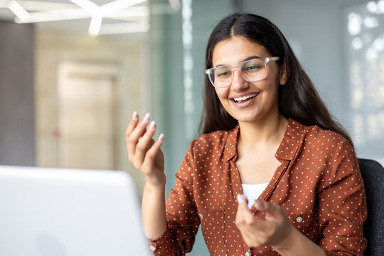 Young indian woman in clear-frame glasses and brown polka-dot shirt smiling and gesturing while engaged in a virtual meeting on her laptop in a bright office setting