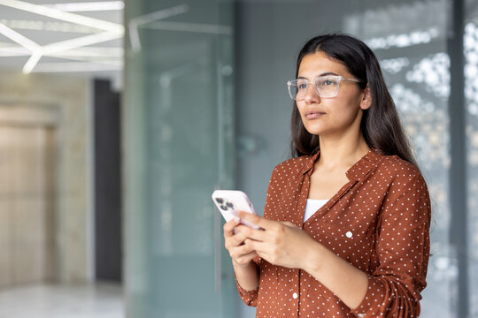 Indian woman wearing glasses and a dotted brown shirt, holding a smartphone and looking away thoughtfully in a contemporary office, representing communication and technology in business