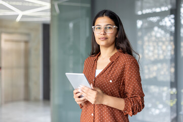 Young indian businesswoman wearing glasses and a polka dot shirt, standing confidently in a modern office, holding a digital tablet and looking at the viewer