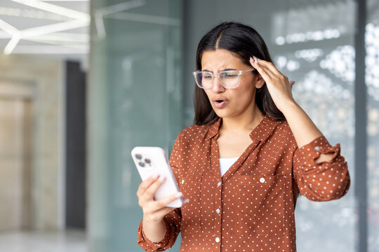 Young woman in a modern office looks shocked and worried as she reads disturbing news on her smartphone, holding the device while processing unexpected bad information