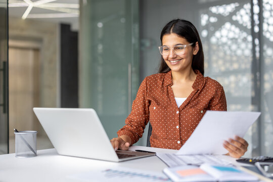 Indian businesswoman wearing glasses sitting at an office desk, smiling while working on a laptop and holding documents, focusing on business, finance, and career growth