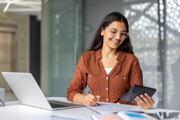Young indian accountant working on documents and using a calculator at her office desk. Managing financial data with a focused yet cheerful expression. Representing successful business and accounting