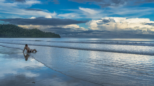 A man sits on the ocean shore at dawn. He leans on his hands. Waves are spreading across the sandy beach. Reflection on wet sand. The sky is blue, the clouds are illuminated. Malaysia. Borneo. 