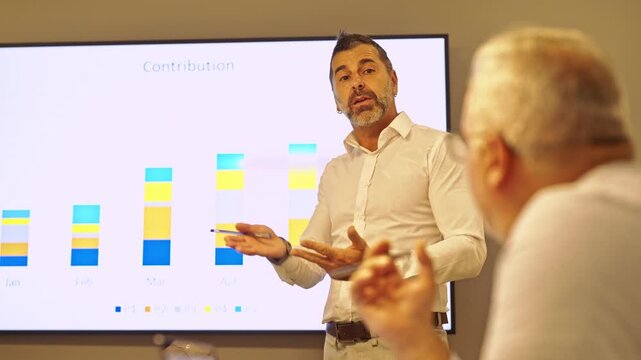 A mature male professional delivers an engaging presentation to a colleague, using a bar chart to illustrate important contribution data.