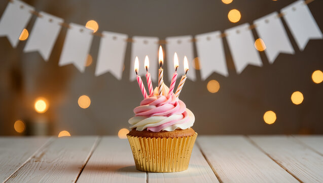 Cupcake with pink and white swirl frosting and five lit birthday candles on top, festive lights and garland in background. Party celebration scene.