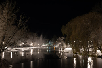A serene winter night scene with a frozen river, glowing streetlights, frosty trees, and a fountain mist reflecting on the calm water.