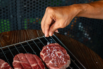Chef hand seasoning raw ribeye steak cuts with coarse sea salt on a stainless steel tray