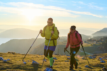 Couple hiking through a grassy mountain trail during sunrise, surrounded by a stunning backdrop of fog in the valleys below. The warm light creates a beautiful atmosphere © Minet