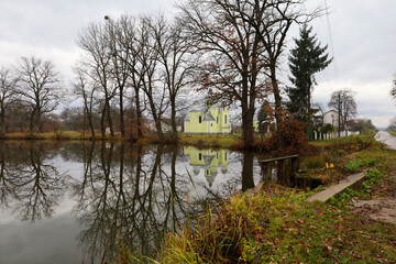 A gloomy autumn landscape featuring leafless trees and a tall spruce on the pond's bank, with a light yellow church building mirrored in the water's surface.