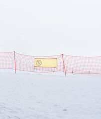 A snowy landscape with a red safety net and a warning sign. The scene is foggy, creating a sense of isolation and caution in winter conditions.