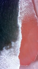 An aerial perspective shows the contrast between the dark blue ocean water and the vibrant pink sand beach as waves gently break.