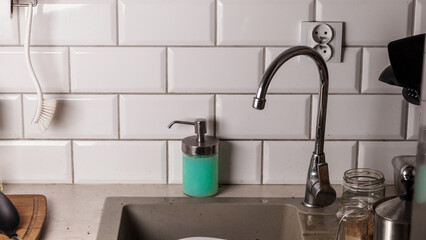 a dirty kitchen sink area with a metal faucet liquid soap dispenser jars and white tile wall creating a realistic home interior setting focused on daily cleaning and household chores