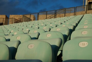 Rows of empty turquoise stadium seats illuminated by golden-hour light. A clean, repetitive pattern ideal for sports, events, architecture, recreation, or venue-related visual concepts.
