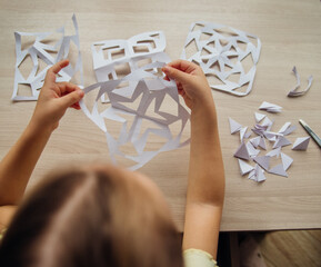 A child creating paper white snowflakes at a table at home. A child cuts patterns on paper with scissors. Decoration for the winter holiday.