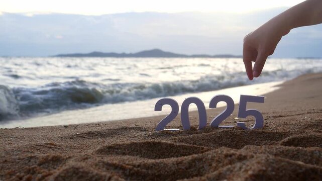 hand picks out the number 5, changing from year 2025 to 2026 for new year sign on summer beach at sunset, with ripple sea wave and beautiful nature at background, 2026 New Year celebration concept