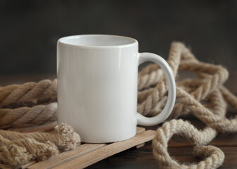 White ceramic coffee mug on dark wood against rustic jute ropes close up, nautical mockup