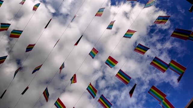 Small LGBT pride flags hanging on strings fluttering in the breeze against a cloudy blue sky. Celebration of gay pride, social acceptance, freedom of love, and human rights