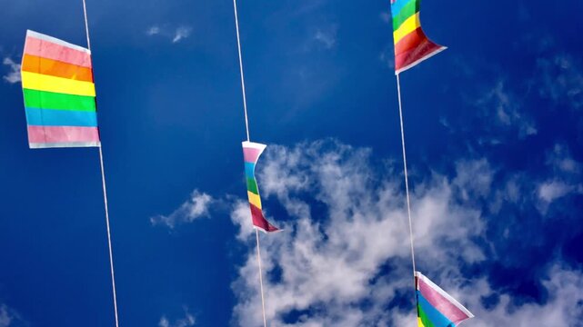 Slow motion view of colorful LGBTQIA plus pride flag garland waving in the wind against a vibrant blue sky with white clouds, symbolizing freedom, love, and community celebration during a sunny day