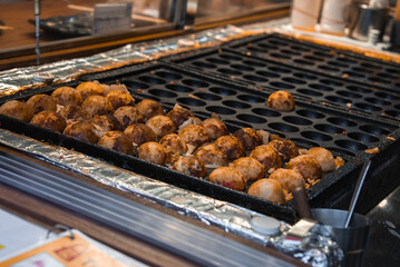 Rows of golden brown takoyaki cook in half sphere molds in Tokyo, with a turning tool, foil lined counter, and a ready to fill section beside crisped snacks