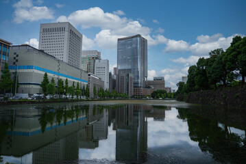 Fototapeta premium Daytime view in central Tokyo shows a canal reflecting glass towers near the Marunouchi district and the Imperial Palace moat, with trees and stone embankment in frame.