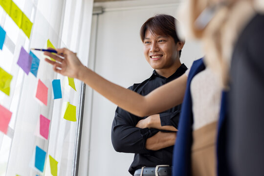 Asian businesspeople are discussing and brainstorming using colorful sticky notes on a glass board in a modern office, working together on a project plan. - Powered by Adobe