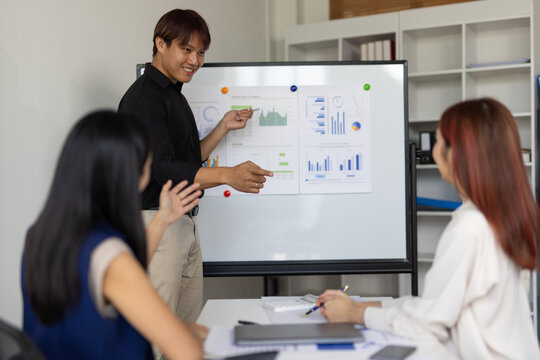 Asian businessman leads a meeting, presenting data and charts on a whiteboard to her colleagues. - Powered by Adobe