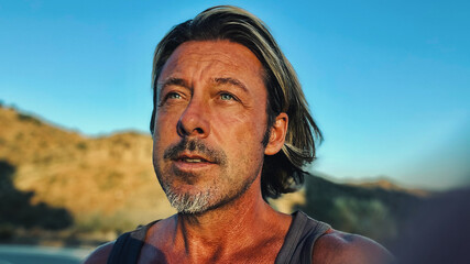 Middle aged tanned man with half long blond hair and grey stubble beard in singlet shirt walking outdoors during golden hour. Hills of Almogia, Malaga, Andalusia, Spain.