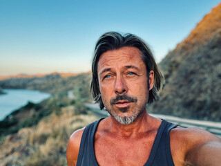 Middle aged tanned man with half long blond hair and grey stubble beard in singlet shirt walking outdoors during golden hour. Hills of Almogia, Malaga, Andalusia, Spain.
