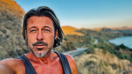 Middle aged tanned man with half long blond hair and grey stubble beard in singlet shirt walking outdoors during golden hour. Hills of Almogia, Malaga, Andalusia, Spain.