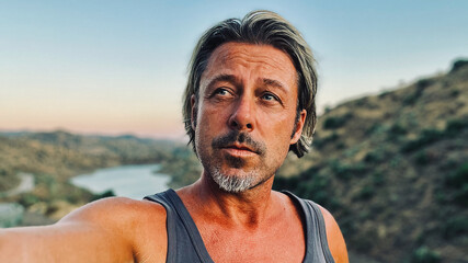 Middle aged tanned man with half long blond hair and grey stubble beard in singlet shirt walking outdoors during golden hour. Hills of Almogia, Malaga, Andalusia, Spain.