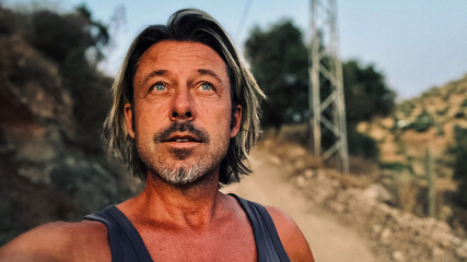 Middle aged tanned man with half long blond hair and grey stubble beard in singlet shirt walking outdoors during golden hour. Hills of Almogia, Malaga, Andalusia, Spain.