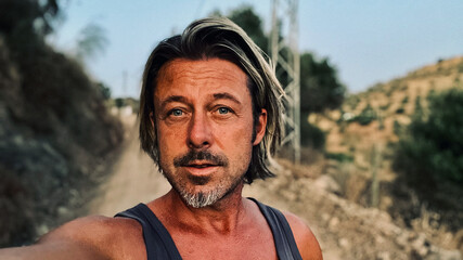 Middle aged tanned man with half long blond hair and grey stubble beard in singlet shirt walking outdoors during golden hour. Hills of Almogia, Malaga, Andalusia, Spain.