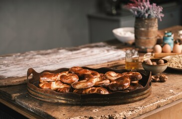 Golden-brown Romanian mucenici sit on a rustic tray in a cozy kitchen. Flour, eggs, and jars filled with ingredients surround them, highlighting the artistry of traditional baking
