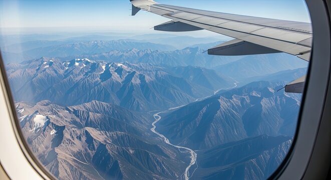 Stunning aerial view from an airplane window looking down on a majestic, snow-capped mountain range with a winding river valley under a clear blue sky