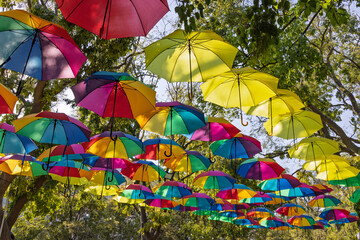 A vibrant canopy of colorful, multi-hued umbrellas suspended over a park in Lisbon, Portugal, creating a cheerful, abstract installation against the trees.