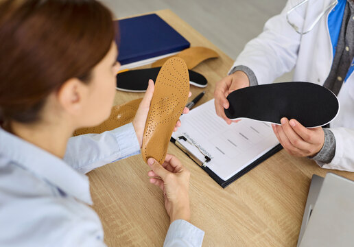 Doctor and patient review orthotics insoles for feet arch support in clinic. Woman and orthopedist compare material of individual orthotic inserts, discuss footwear and foot health choice at desk