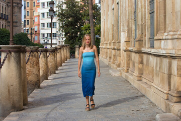 A young, beautiful blonde woman in a blue dress walks between stone columns. The woman is on...