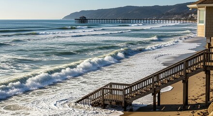 A scenic coastal landscape with a wooden staircase leading down to a sandy beach, with ocean waves, a long pier, and distant hills under a clear sunny sky