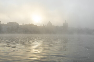 Fototapeta premium Vltava river flowing past historic prague buildings and charles bridge during a foggy sunrise, creating an evanescent atmosphere