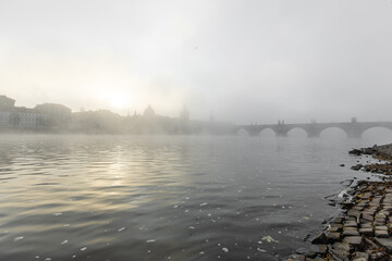 Vltava river flowing past charles bridge and prague cityscape, dramatically obscured by thick morning fog and soft light