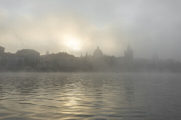 Naklejka premium Prague charles bridge appearing as silhouettes across the vltava river, bathed in the soft glow of a hazy sunrise