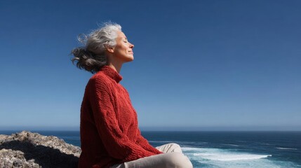 Woman sitting on a rocky cliff overlooking the ocean. she is wearing a red sweater and has her hair tied up in a bun. the woman is looking up towards the sky with a peaceful expression on her face.