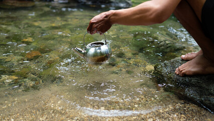 Person Pouring Water From Teapot Into Clear Stream While Kneeling By Rocks Near Forest Shore