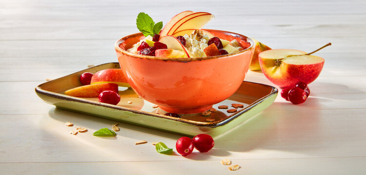 Koerniger Frischkaese mit frischem Apfel und getrocknetetn Fruechten in ornagener Steingutschale auf hellem Holzfond, M&uuml;sli, Apfel, Fr&uuml;hst&uuml;ck, Cranberry, Fr&uuml;chte, 