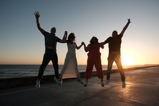 Four friends or family of four are jumping together at sunset near the ocean, holding hands and celebrating a joyful moment in silhouette.