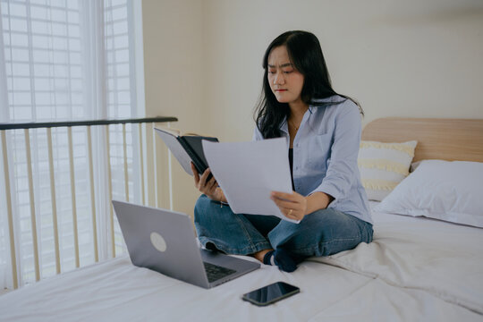 Focused asian woman works on financial documents, sorting reports and analyzing data at home, sitting on bed in cozy bedroom interior. Accounting and finance concept. - Powered by Adobe