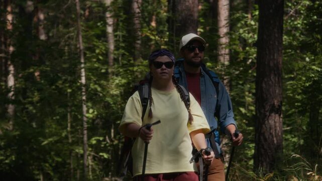 Man and young woman with down syndrome, wearing sunglasses, backpacks, and using hiking poles, hiking through sun-dappled forest