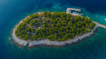 Small, verdant island with a rocky coastline, a pier, and boats rests like a jewel in the middle of...