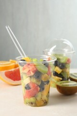 Tasty fruit salad in plastic cups and ingredients on beige table, closeup. Healthy snack